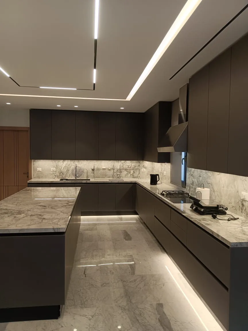 KZ kitchen Ghazze, Beqaa Lebanon — wide view of dark brown handleless cabinetry with Super White quartzite worktops and backsplash, plinth LED strip lighting, recessed linear ceiling lights and oversized waterfall island on marble floor — KITWOOD Lebanon completed project
