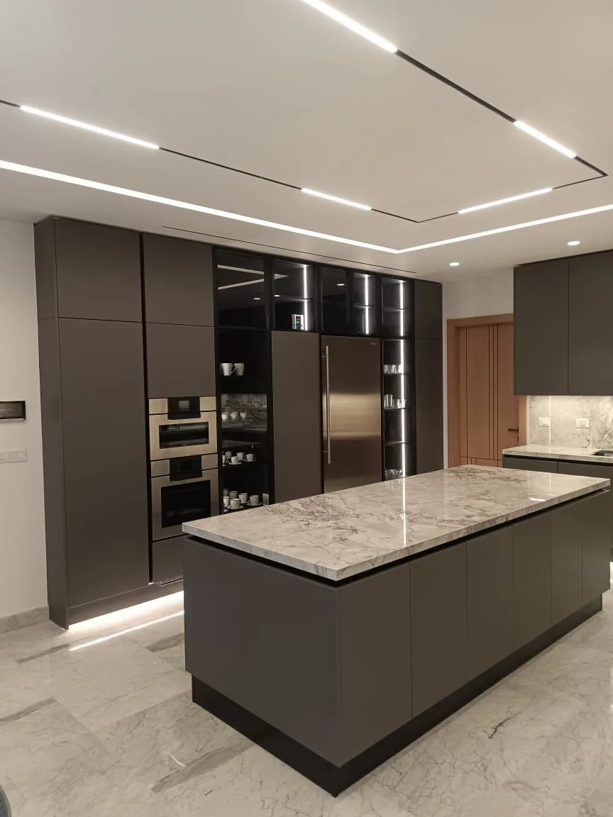 KZ kitchen Ghazze, Beqaa Lebanon — full-height dark brown tall unit run with built-in oven and combi-microwave column, smoked glass display cabinets, integrated American-style stainless steel fridge-freezer and matching handleless island with quartzite top — KITWOOD Lebanon completed project