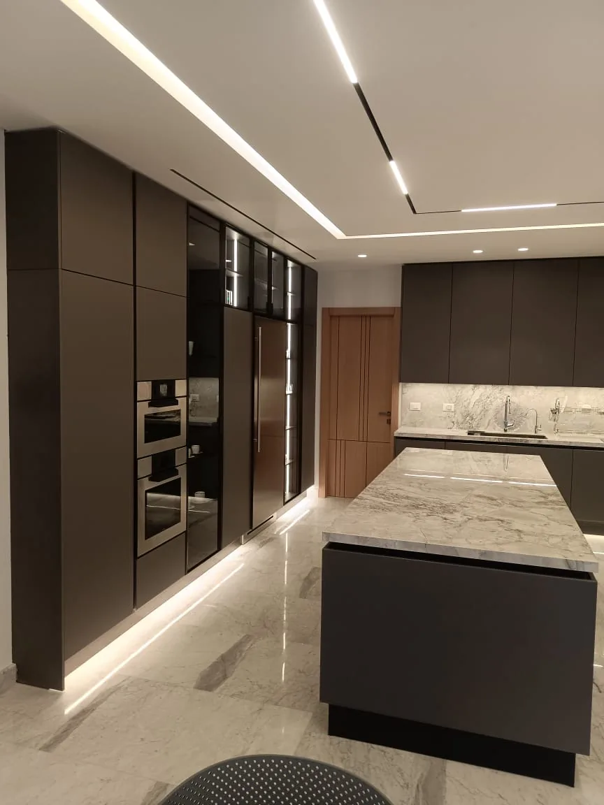 KZ kitchen Ghazze, Beqaa Lebanon — angled view showing full-height dark brown tall units with appliance column and smoked glass cabinets, oak service door, quartzite waterfall island and quartzite backsplash with under-cabinet and plinth LED lighting on polished marble floor — KITWOOD Lebanon completed project