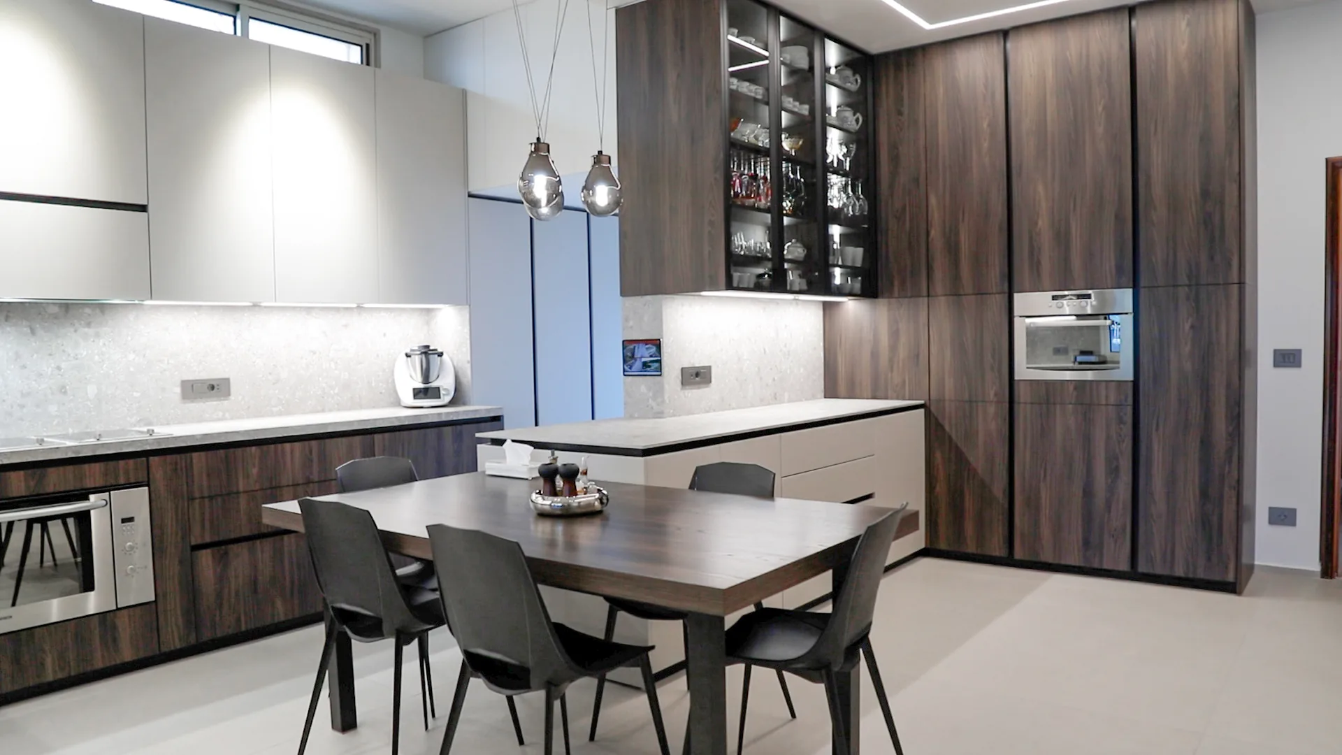 DV kitchen Dbayeh, Metn Lebanon — view of dark walnut full-height tall units with built-in oven and combi-microwave column, illuminated walnut and glass stemware display cabinet, white handleless wall units, terrazzo backsplash, walnut dining table with smoked-glass pendant lights — KITWOOD Lebanon completed project