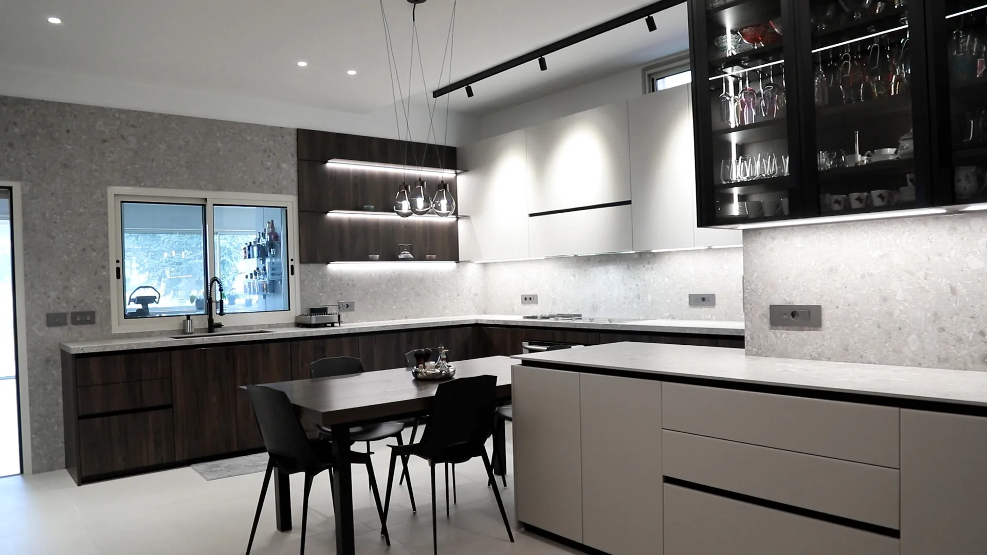 DV kitchen Dbayeh, Metn Lebanon — wide view of central white handleless island with terrazzo top, dark walnut base cabinetry with LED-lit open shelving, smoked-glass display cabinet wall for stemware and dining table with cluster pendant lights — KITWOOD Lebanon completed project