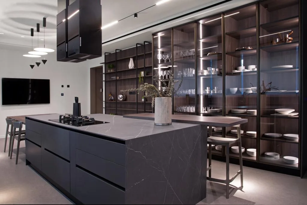 YK kitchen Yarze, Baabda Lebanon — focused view of Pietra Grey marble waterfall island with integrated gas hob and breakfast bar, charcoal handleless base cabinetry, full-height bronze-frame walnut and glass display cabinet wall with LED-lit shelving for stemware and crockery and dark conical extractor hoods — KITWOOD Lebanon completed project