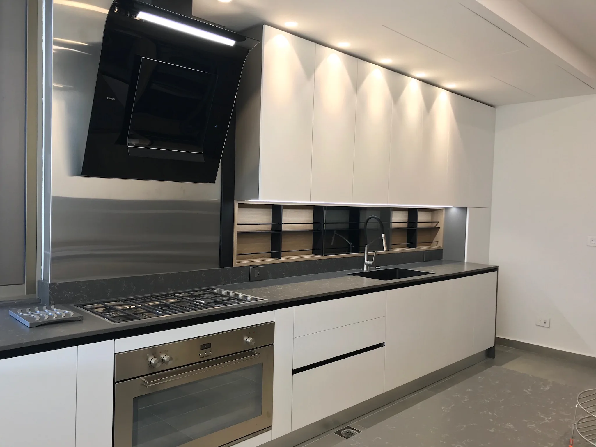 BL kitchen executed in Badaro, Beirut Lebanon — minimalist single-wall white handleless kitchen with grey stone worktop, sculptural curved black extractor hood, integrated stainless gas hob and oven, recessed oak open shelving niche with black metal frame and recessed ceiling downlights — KITWOOD Lebanon completed project