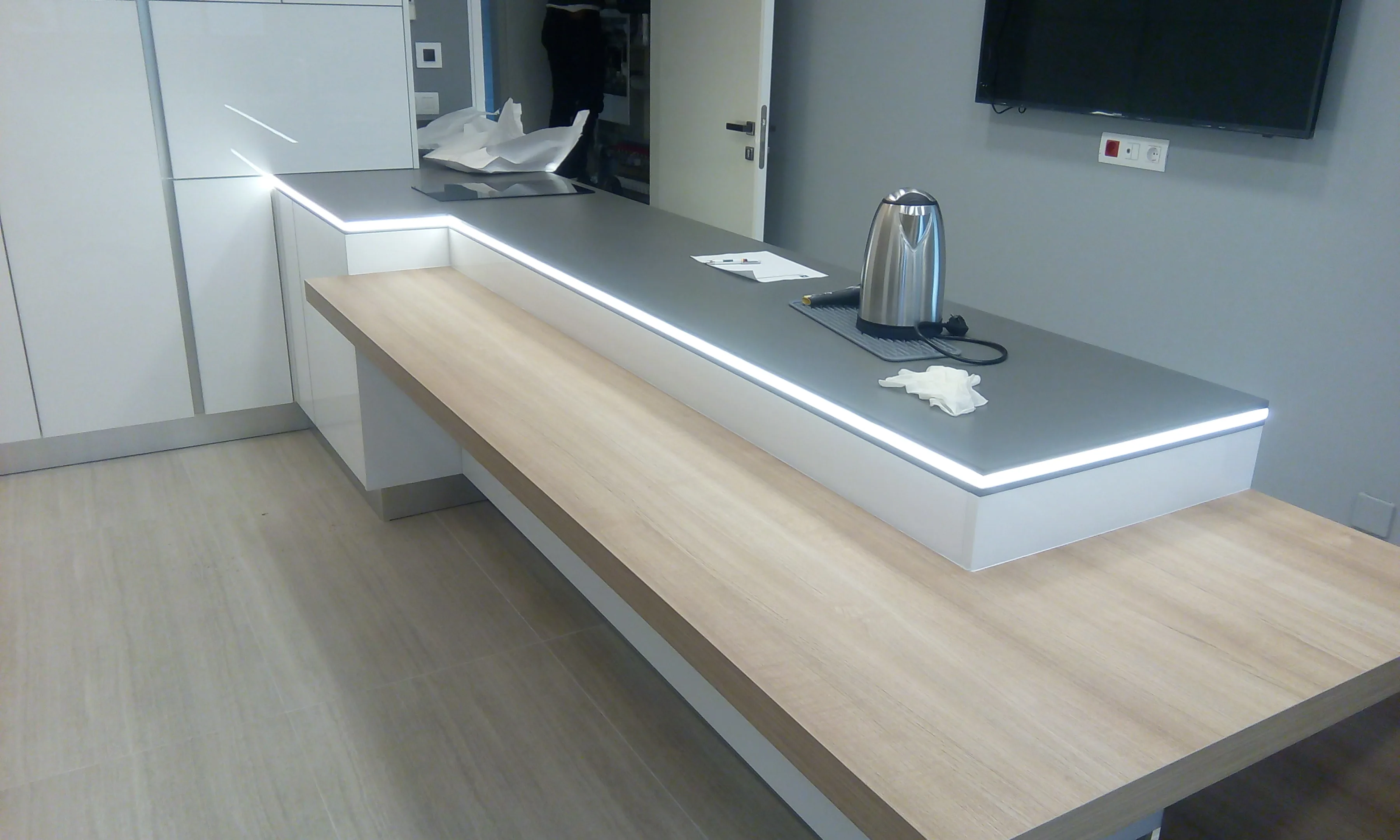 SF kitchen executed in Sin el Fil, Metn Lebanon — compact white handleless kitchen with grey stone worktop and integrated LED edge lighting, oak breakfast-bar extension, integrated induction hob and wall-mounted television above — KITWOOD Lebanon completed project