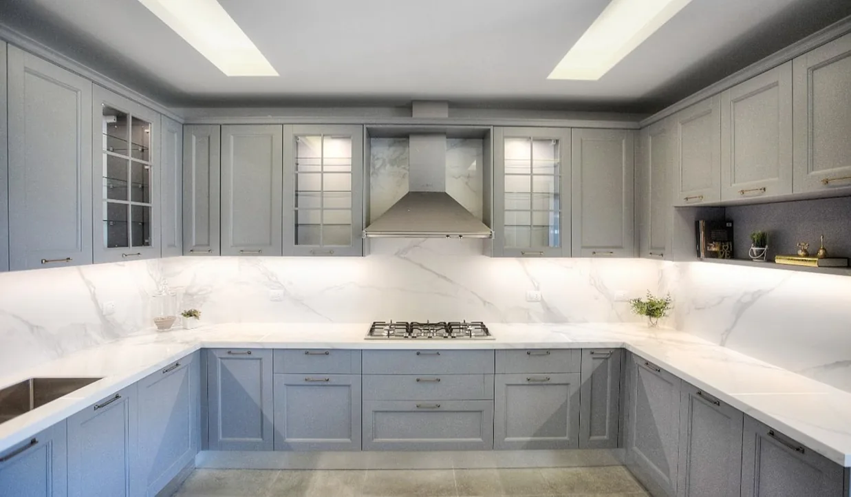 BM kitchen executed in Broummana, Metn Lebanon — classic grey-blue shaker U-shape kitchen with Calacatta marble countertops and full-height marble backsplash, glazed wall cabinets with brass handles, central stainless extractor hood, integrated gas hob and recessed ceiling light coves — KITWOOD Lebanon completed project