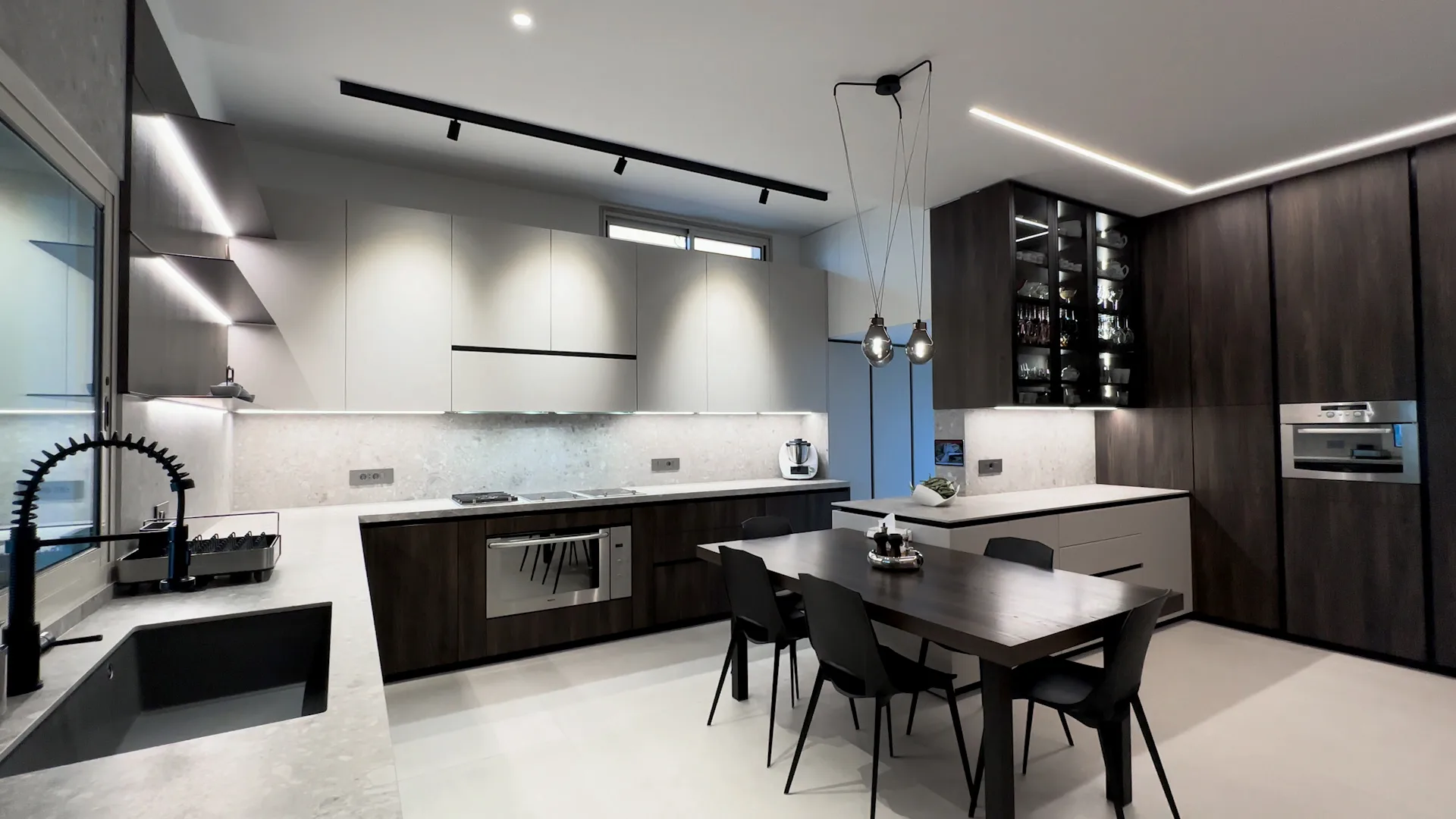 MR kitchen executed in Rabweh, Metn Lebanon — open-plan kitchen with dark walnut veneer base cabinets and full-height walnut tall unit run with integrated stainless steel oven column, cream matte handleless wall units, speckled cream terrazzo worktops and full-height backsplash, walnut LED-lit floating open shelves and smoked-glass LED-lit walnut stemware display cabinet with black metal frame, professional black gooseneck tap, walnut dining table with black chairs under cluster of smoked-glass bulb pendant lights and dedicated walnut prep kitchen / scullery with matching terrazzo — KITWOOD Lebanon completed project