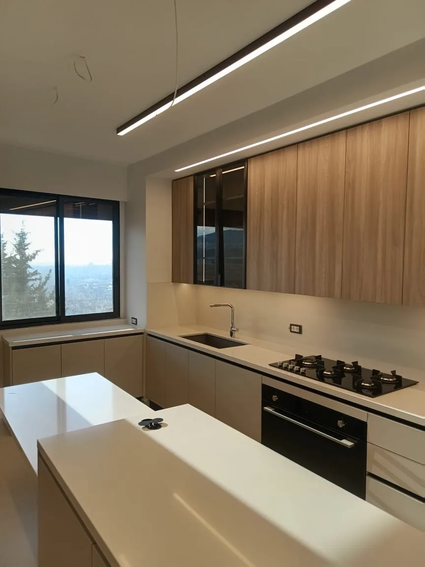 TS kitchen executed in Beit Mery, Metn Lebanon — minimalist handleless kitchen with light oak vertical-grain veneer wall units, smoked-glass corner display cabinet with black metal frame, beige matte handleless base cabinetry, white quartz worktops and waterfall island, integrated black gas hob and built-in oven column, single suspended linear LED bar and black-framed picture window overlooking the Mount Lebanon valley — KITWOOD Lebanon completed project