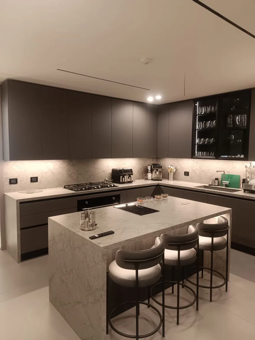 GH kitchen executed in Aïn Saadeh, Metn Lebanon — L-shape handleless kitchen in dark taupe matte lacquer with grey-veined marble worktops and full-height matching marble backsplash, smoked-glass LED-lit corner display cabinet for stemware, integrated black gas hob, induction hob, combi-oven column and undermount sink with chrome tap, cantilevered marble waterfall island with bar overhang and three round cream-leather bar stools on light porcelain flooring — KITWOOD Lebanon completed project