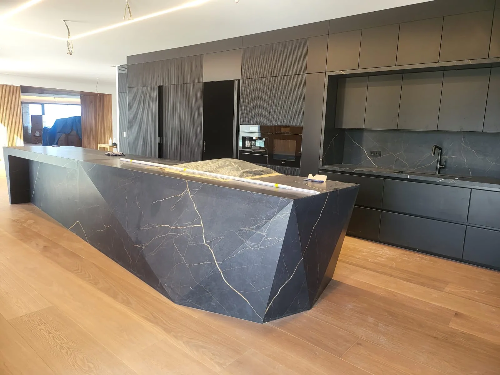 VL kitchen executed in Saifi Village, Beirut Lebanon — flagship architectural installation with sculptural faceted geometric Pietra Grey marble waterfall island showcasing dramatic gold and amber veining, charcoal matte handleless base cabinets with matching Pietra Grey worktops and backsplash, full-height reeded vertical-fluted dark grey tall units with integrated dark appliance column and natural light oak plank flooring — KITWOOD Lebanon completed project
