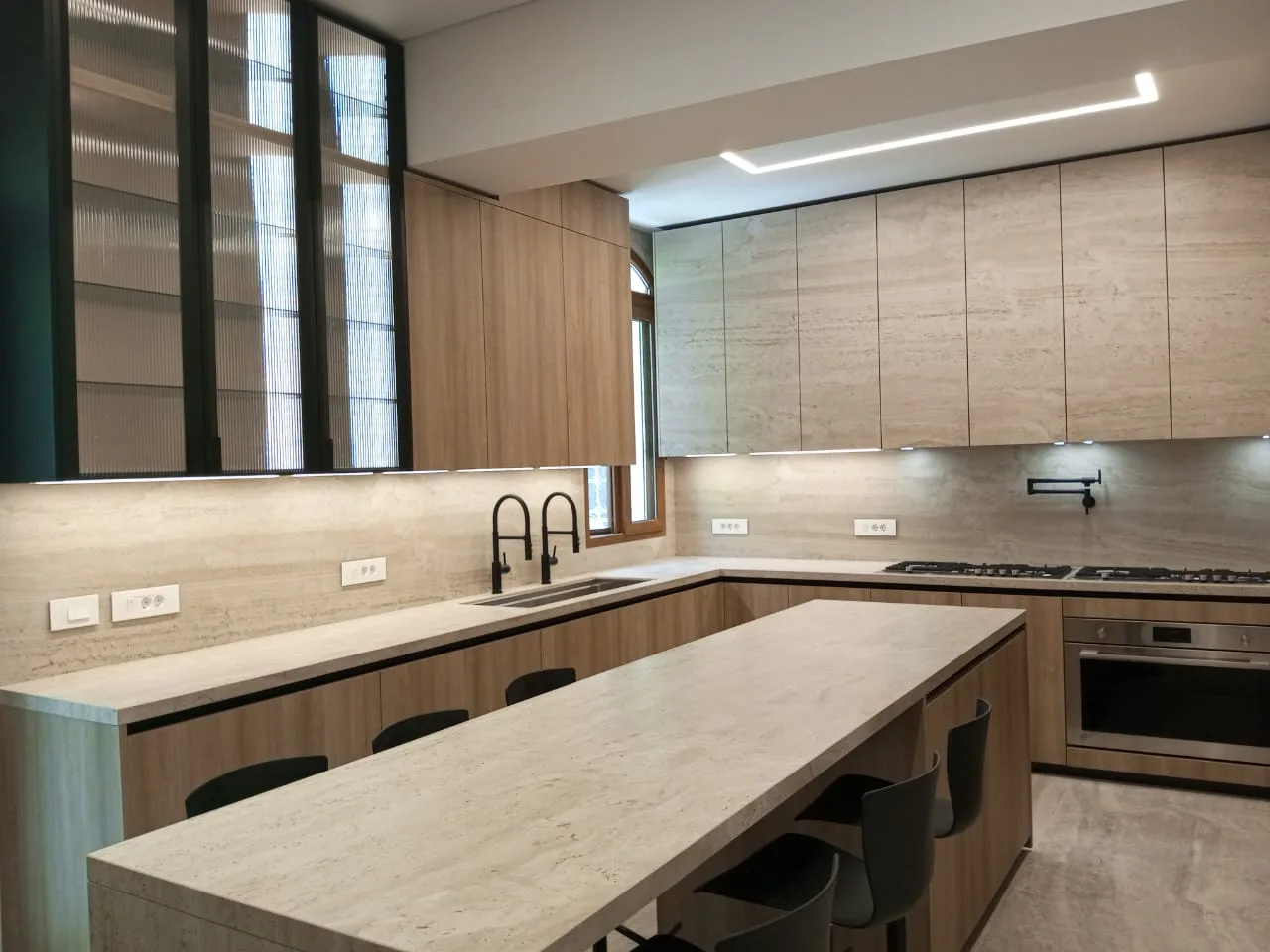 AD kitchen executed in Adma, Kesrwan Lebanon — U-shape villa kitchen with light oak veneer handleless base and full-height tall units, travertine-fronted wall units with full-height travertine backsplash and worktops, tall reeded fluted-glass display cabinets with black metal frames, twin stainless steel American-style fridge-freezers flanked by oak appliance columns each housing built-in oven and combi-microwave, professional black pot-filler and gooseneck taps, integrated gas hob and stainless steel oven and long travertine peninsula dining bar with black moulded bar stools on black pedestal bases and sculptural dark moulded chairs — KITWOOD Lebanon completed project