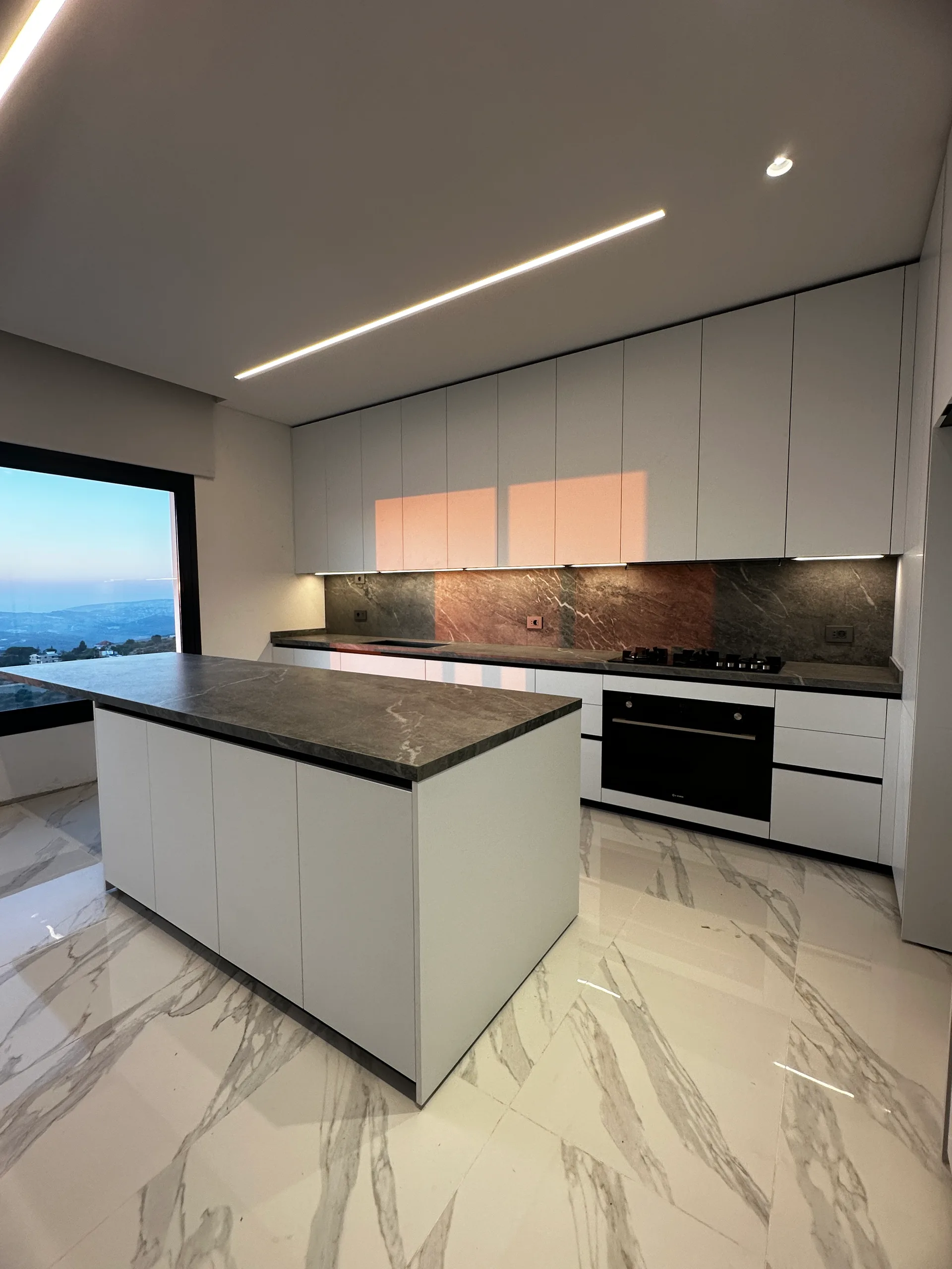 MS kitchen executed in Bhamdoun, Mount Lebanon — minimalist white handleless single-wall kitchen with dramatic bronze and copper-veined dark stone backsplash and matching island waterfall worktop, white matte tall units and base cabinets, integrated black gas hob and built-in oven, single suspended linear LED bar and floor-to-ceiling picture window overlooking Mount Lebanon, on polished Calacatta marble porcelain flooring with bold grey veining — KITWOOD Lebanon completed project