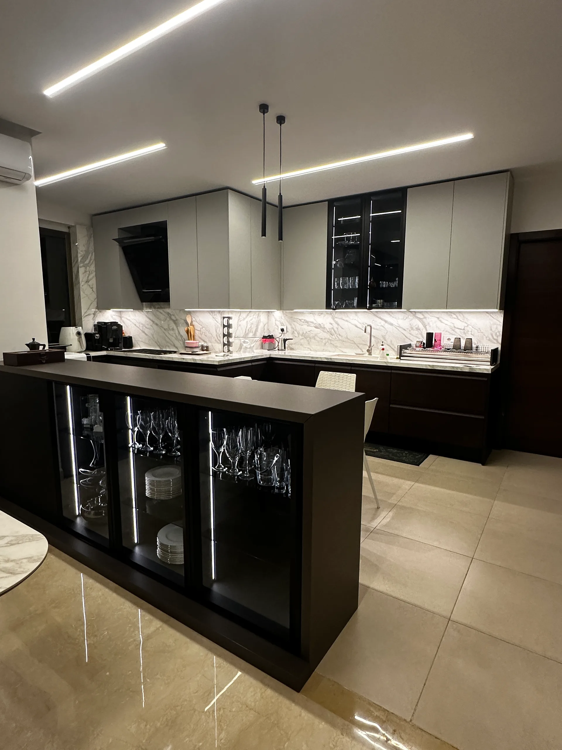 EM kitchen executed in Mansourieh, Metn Lebanon — U-shape handleless kitchen with dark espresso wenge base cabinetry, light grey matte handleless wall units, Calacatta marble worktops and full-height marble backsplash, angled sculptural light grey extractor hood, LED-lit smoked-glass stemware display cabinets, twin slim suspended cylinder pendant lights and espresso bar island with three glass-front LED-lit display panels for stemware and dinnerware on polished cream travertine flooring — KITWOOD Lebanon completed project
