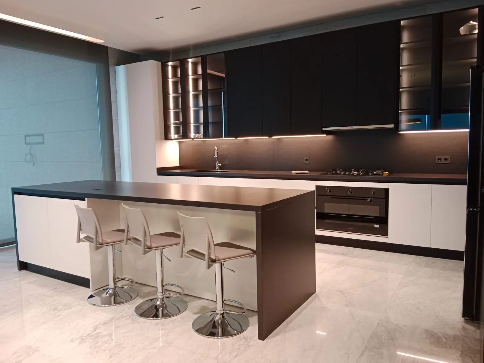 EH kitchen executed in Elissar, Metn Lebanon — long galley kitchen with floor-to-ceiling dark espresso wenge wall and base cabinetry, integrated full-height fridge column, built-in walnut LED-lit open display niche with cantilevered shelves, dark espresso worktops with integrated gas hob and built-in oven, central island with cream matte base and matching espresso top and three cream-leather bar stools on chrome bases over polished Calacatta marble flooring — KITWOOD Lebanon completed project