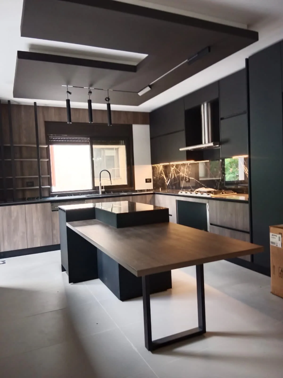 KH kitchen executed in Kornet El Hamra, Metn Lebanon — architectural kitchen with dramatic stepped dark grey dropped ceiling soffit and exposed black track lighting, anthracite matte handleless tall and wall units, full-height light oak veneer open shelving niche with black metal frames, exotic dark stone backsplash with bold gold and amber veining, stainless steel chimney hood, central anthracite island in dark stone with perpendicular extending walnut dining table on black steel U-base and cluster of black cylindrical pendant lights — KITWOOD Lebanon completed project