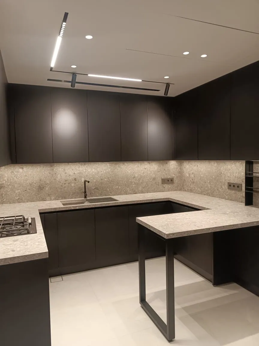 Residence J.B. kitchen executed in Downtown Beirut, Lebanon — minimalist U-shape handleless kitchen with floor-to-ceiling anthracite matt lacquer tall and wall units, light grey terrazzo stone worktop and full-height backsplash, integrated stainless steel gas hob, undermount sink with brushed bronze mixer tap, peninsula breakfast bar with terrazzo top on slim black steel U-frame, suspended linear LED track lighting bar and large-format off-white porcelain flooring — KITWOOD Lebanon completed project